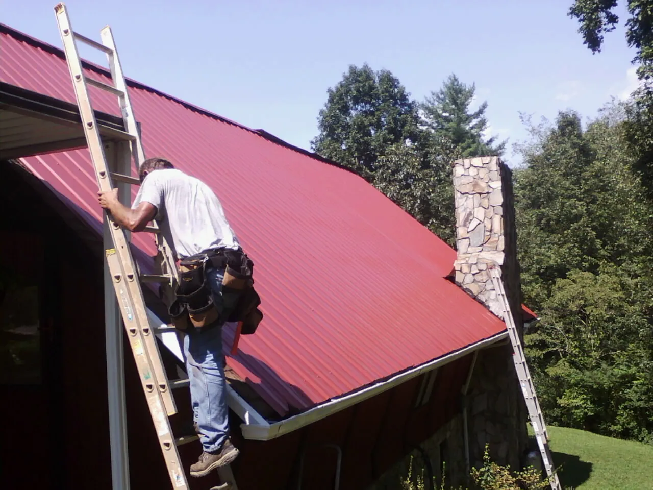 Roofer on a ladder beside a red metal roof and stone chimney — Soffit & Fascia Repair by Roofing Brothers in Leipsic